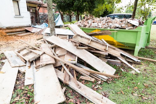 The image shows a collection of discarded wooden planks and broken panels, some with painted or weathered surfaces, piled haphazardly on a grassy area next to a large green skip filled with a mixture of rubble and debris. The wooden materials vary in size and shape, with some pieces leaning against each other and others lying flat. In the background, there are trees with green foliage and a residential building partially visible, indicating an outdoor garden or yard setting. The scene appears to be part of a garden clearance process, consistent with rubbish removal services in Purley. The lighting suggests daytime with natural sunlight casting soft shadows, highlighting the textures of the old wood and the surrounding environment. This image exemplifies typical waste removal tasks handled by Garden Clearance Purley, focusing on the clearance of old wooden garden structures or fencing materials.