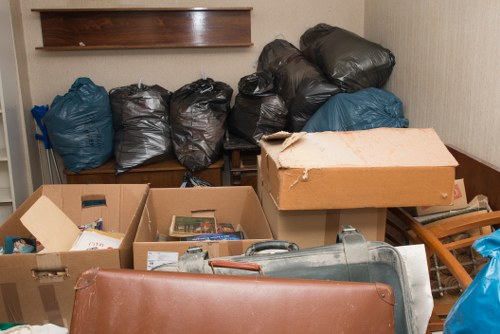 The image shows a small indoor space, possibly a storage room or garage, filled with various rubbish items. In the foreground, there are open cardboard boxes containing miscellaneous waste items such as paper, packaging, and small household objects. To the right, a partially crushed cardboard box covers a metallic cylindrical object that appears to be a vacuum cleaner or similar device. Behind these boxes, several black and blue plastic rubbish bags are stacked on a wooden shelf or bench, with the black bags appearing to contain general waste and the blue bags possibly holding recyclable or garden waste. The bags are slightly bulging, indicating they are filled with rubbish. The environment is well-lit, with neutral-colored walls, and the space appears to be somewhat cluttered, typical of an area awaiting rubbish collection or clearance. The image aligns with services offered by Garden Clearance Purley, which handles rubbish removal in the Purley area, including disposal and clearance of household and garden waste.