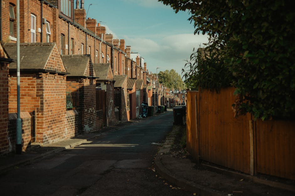 A narrow residential street lined with terraced brick houses featuring small front gardens and sloped tiled roofs. The street has a paved, slightly worn surface with a few visible cracks and patches. 