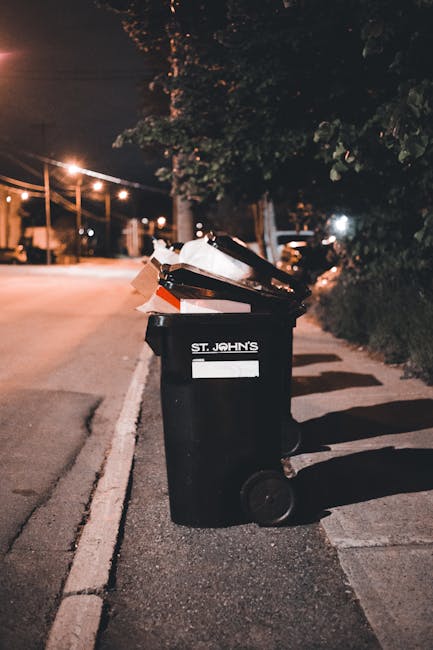 A person in a dark-colored long-sleeve garment hands a crumpled brown paper bag to another individual dressed in a light gray sweater, as part of a rubbish collection process outside a building. The s